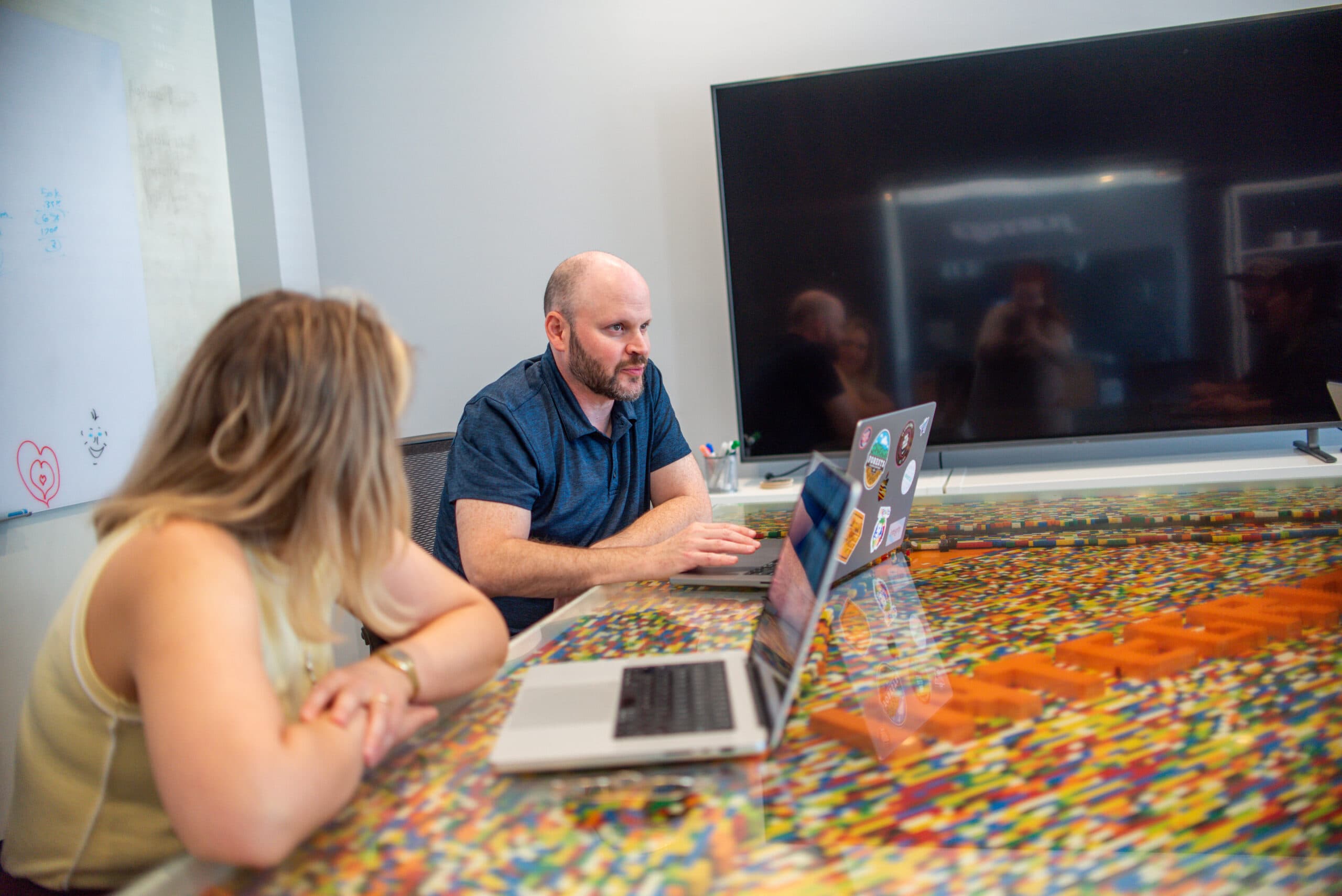 Two team members working on laptops at a table inlaid with colorful toy bricks